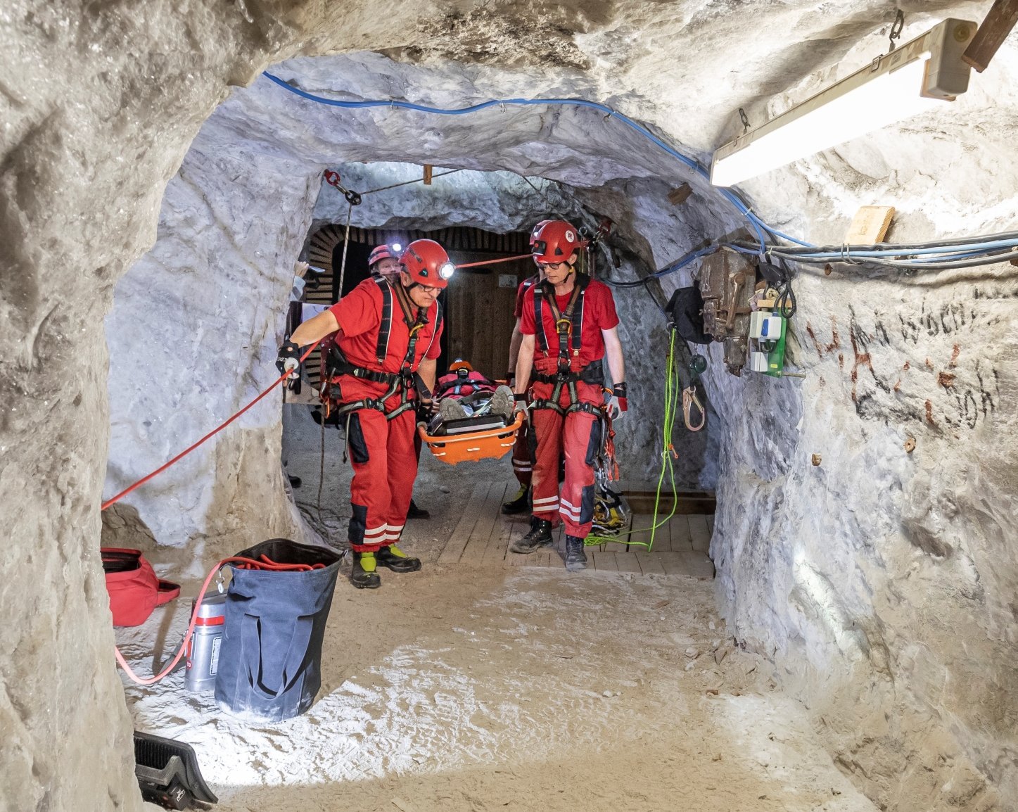 Zwei Männer in roten Arbeitsanzügen gehen durch einen Bergwerkschacht. Zwischen ihnen tragen sie eine Trage mit einem Verletzen.