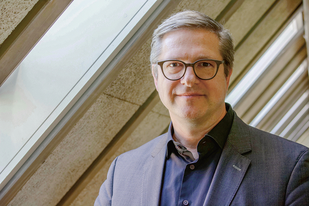 Kommunikationswissenschaftler Frank Brettschneider Porträtfoto Frank Brettschneider. Mann mit Brille. Er trägt ein dunkelblaues Hemd und eine Jacket und steht unter einer Dachschräge.